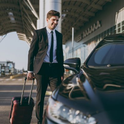 Full size bottom view young traveler businessman young man in black dinner suit going walk outside at international airport terminal with suitcase to car booking taxi Air flight business trip concept.